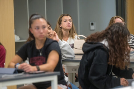A woman seated in a classroom looks toward the front of a room while a group around her are also listening. The group is wearing Lafayette shirts.