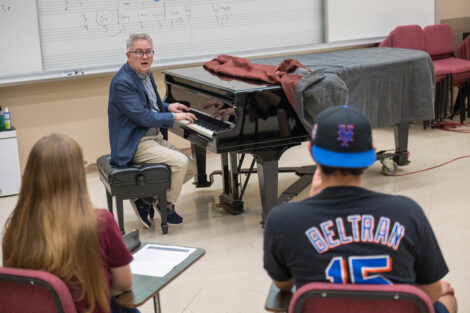 A professor plays the piano while two individuals watch in a classroom.