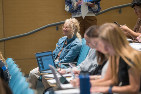 A woman seated in a classroom smiles. She is wearing a jean jacket.
