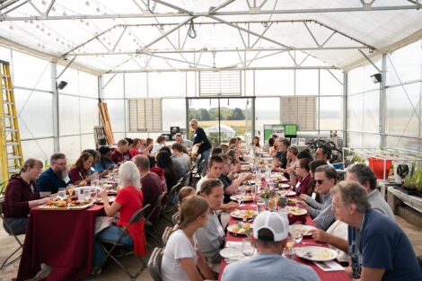 Attendees at the LaFarm to Table breakfast are seated at long tables with maroon tablecloths in the greenhouse.