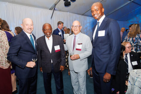 Four alumni stand and smile at the camera. They are all wearing suits.