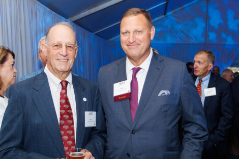 Two gentlemen smile at the camera. They are both wearing gray suits and maroon ties.