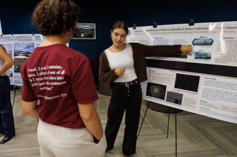 A student points to their research project while a faculty member looks on.