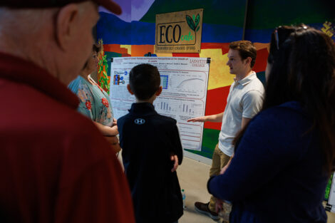 A student stands in front of their research poster, talking to families about their discoveries.
