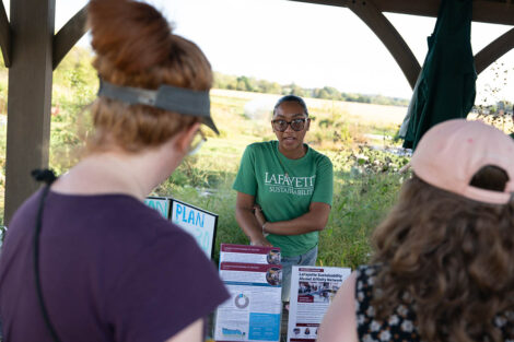 A person in a Lafayette Sustainability shirt stands behind a table holding sustainability posters, speaking to attendees.