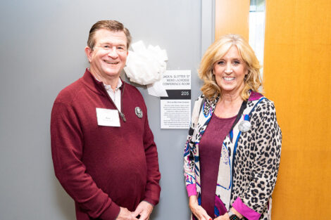 Harry Dombroski and Jan Ellen Slotter Dombroski stand in front of the men's lacrosse conference room. There is a white bow next to a plaque.