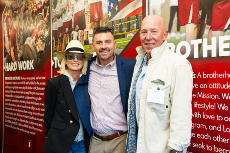 Cindy ’80, P’13’18 and Jud Linville '79 P’13 ‘18 with Coach Pat Myers stand in the men's lacrosse locker room in the Center. Cindy is wearing a hat and sunglasses.