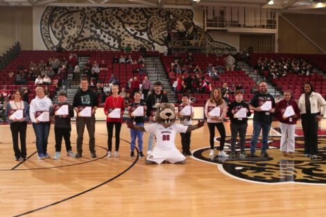 Roary the Leopard mascot kneels in the middle of Kirby Sports Center, surrounded by 13 Easton area students and teachers holding certificates.