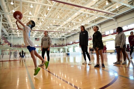 A girl jumps in mid-air to shoot a basketball as others watch.