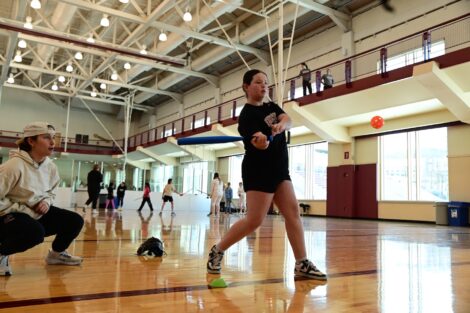 A young participant swings a bat as a softball player looks on.