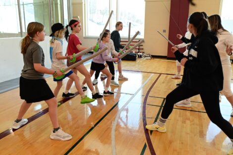 Members of women's fencing team and young participants face each other with foam swords.