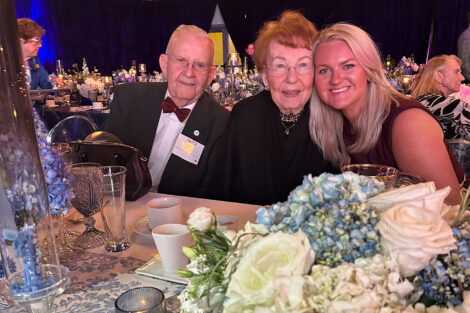A family of three are seated at a table at a dinner. there are blue flowers in the foreground. A man is wearing a maroon bow tie.