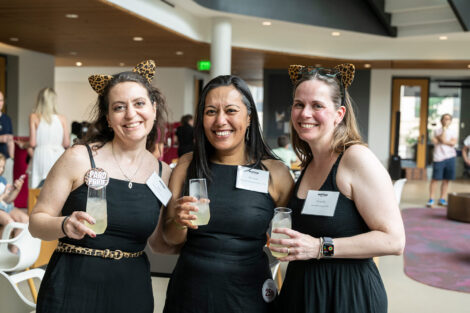 Three women are holding drinks and standing and smiling.