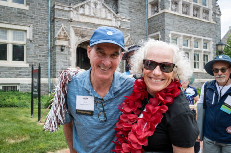 Two alumni are in front of Hogg Hall. One is wearing a maroon flowered lei.