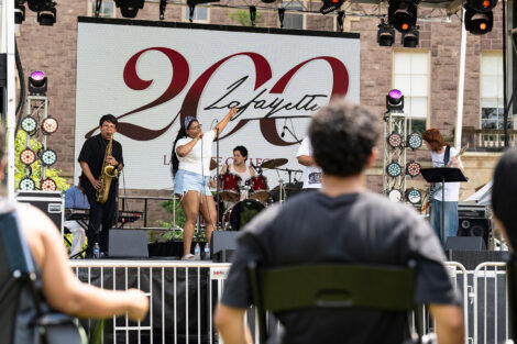 A band plays on a stage on the Quad. The lead singer is wearing a bandana on her head.