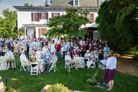 A large group of alumni mingle on the President's House back lawn. President Hurd stands at a podium.