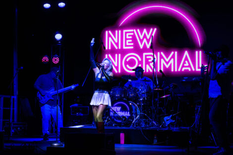 The band New Normal sings on stage. There is a New Normal pink neon sign behind the lead singer.