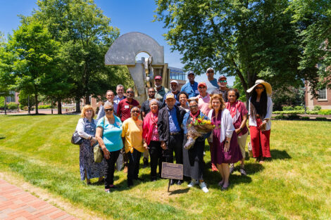 A large group of alumni stand in front of the Transcendence statue. One alumna is holding flowers. President Hurd is in the photo.