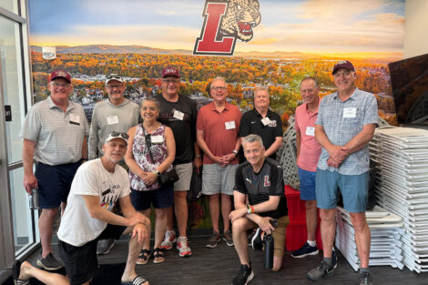 A group of alumni pose in front of a wall with an aerial image of campus. The Lafayette athletics logo is in the background.