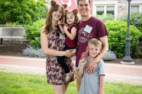A family of four is standing and smiling at the camera. A young boy holds up a maroon foam finger.