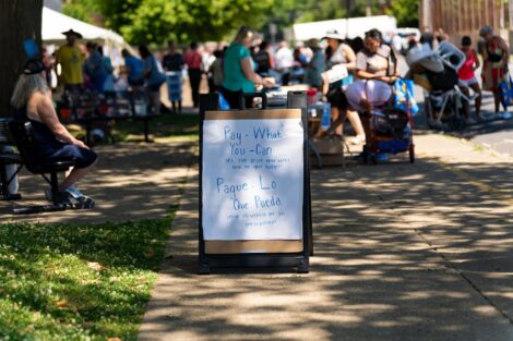 An A-frame sign reads Pay what you can, with shoppers in the background visiting the West Ward Sale.